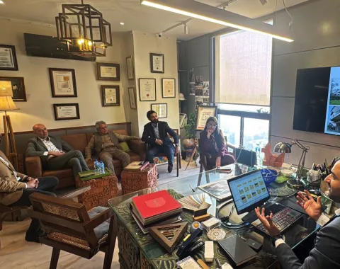 Ein Büro mit mehreren Personen, die in einem Meeting sitzen. Eine Person sitzt an einem Schreibtisch mit einem Laptop, während andere auf Stühlen und einem Sofa Platz genommen haben. An der Wand hängen Zertifikate und ein Bildschirm zeigt architektonische Entwürfe.