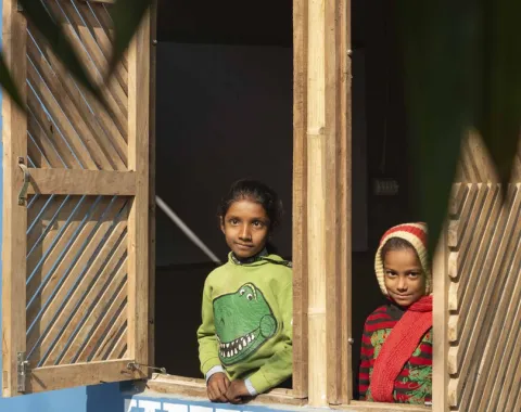 Zwei Kinder schauen aus einem offenen Fenster eines Gebäudes mit blauer Wand und dekorativem Muster. Das Fenster hat hölzerne Fensterläden. Im Vordergrund sind unscharfe Blätter zu sehen.