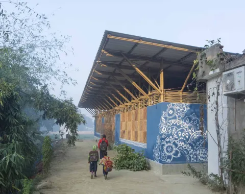 Ein modernes Gebäude mit einem Dach aus Bambus und Wellblech, verziert mit blauen, traditionellen Mustern. Im Vordergrund gehen mehrere Personen auf einem sandigen Weg entlang, umgeben von grüner Vegetation.