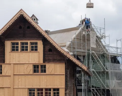 Ein traditionelles Gebäude wird renoviert. Links ist eine Holzfassade zu sehen, rechts stehen Gerüste mit Bauarbeitern auf dem Dach. Der Himmel ist bewölkt.