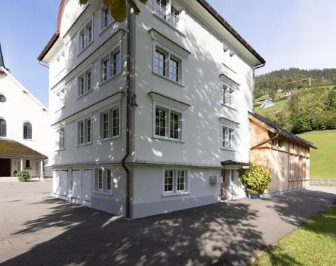 Ein weißes, mehrstöckiges Gebäude mit traditionellen Fenstern steht neben einer Kirche im ländlichen St. Galler Rheintal. Im Hintergrund sind grüne Hügel zu sehen. Der Schatten eines Baumes fällt auf den gepflasterten Vorplatz.