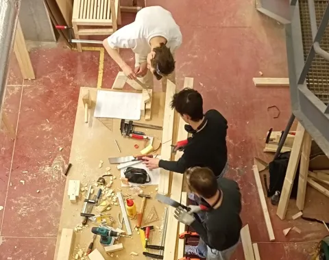 Vier Studierende arbeiten gemeinsam in einer Werkstatt an einem Holztisch. Sie sind von oben zu sehen und verwenden verschiedene Werkzeuge, um ein Möbelstück zu bauen. Holzstücke und Werkzeuge liegen auf dem Tisch verteilt.