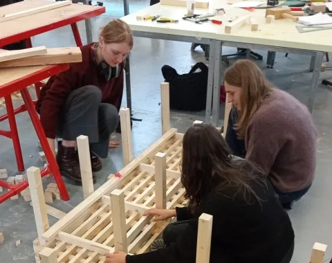 Drei Studierende arbeiten gemeinsam an einem Holzprojekt in einer Werkstatt. Sie montieren ein Möbelstück aus Holzlatten. Im Hintergrund sind Werkbänke und Werkzeuge sichtbar.