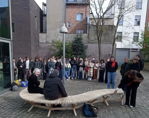 Eine Gruppe von Studierenden steht im Freien um eine geschwungene Holzbank, die von zwei Personen genutzt wird. Die Szene findet auf einem gepflasterten Platz vor modernen Gebäuden statt.