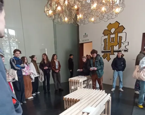 Eine Gruppe von Studierenden steht in einem Raum der Universität Antwerpen um ein modernes, aus Holzlatten gefertigtes Möbelstück. Über ihnen hängt eine große, kunstvolle Lampe. Im Hintergrund ist ein Wandlogo der Universität zu sehen.