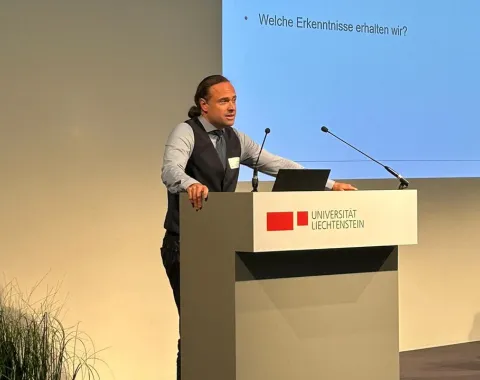 Ein Redner steht an einem Podium mit dem Logo der Universität Liechtenstein. Im Hintergrund ist eine Präsentationsfolie mit dem Titel "Reflexion" und den Fragen "Wo stehen wir?", "Was betrachten wir?" und "Welche Erkenntnisse erhalten wir?" zu sehen. Links im Bild sind Pflanzen dekorativ platziert.