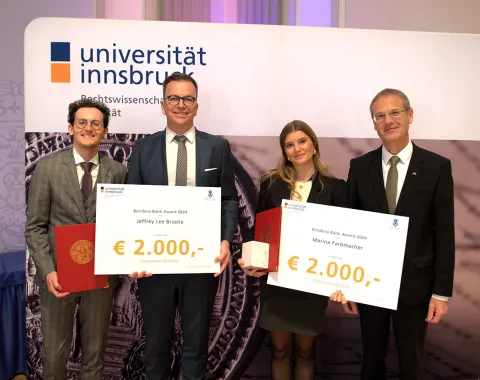 Vier Personen stehen nebeneinander und halten große Schecks mit der Aufschrift "Bendura Bank Award 2024" und einem Betrag von 2.000 Euro. Im Hintergrund ist ein Banner der Universität Innsbruck zu sehen. Die Personen sind formell gekleidet und lächeln in die Kamera.