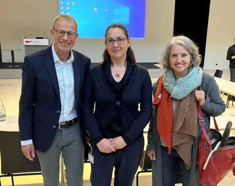 Drei Personen stehen lächelnd in einem Auditorium der Universität Liechtenstein. Im Hintergrund sind ein Rednerpult und eine Leinwand mit einer Präsentation zu sehen.