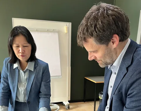 Zwei Personen in formeller Kleidung betrachten Bücher auf einem Tisch in einem Konferenzraum. Im Hintergrund ist ein Flipchart zu sehen.