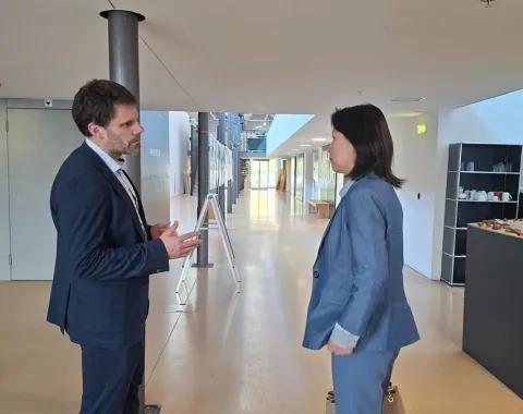 Zwei Personen in einem modernen Gebäude im Gespräch. Beide tragen Business-Kleidung. Im Hintergrund sind helle Flure und moderne Einrichtungselemente zu sehen.