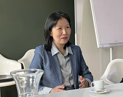 Eine Person in einem blauen Blazer sitzt an einem Konferenztisch mit einer Tasse Kaffee und einem Notizblock. Im Hintergrund ist ein Flipchart zu sehen.