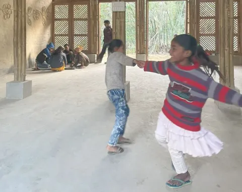 Kinder spielen und tanzen in einem neu gebauten Schulgebäude aus Bambus in Bihar, Indien. Im Hintergrund sitzen weitere Kinder auf dem Boden. Das Gebäude hat eine natürliche, lichtdurchflutete Atmosphäre.