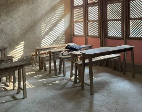 Ein einfacher Klassenraum mit Holztischen und -bänken, durchflutet von Sonnenlicht, das durch Fenster mit Holzlamellen fällt.