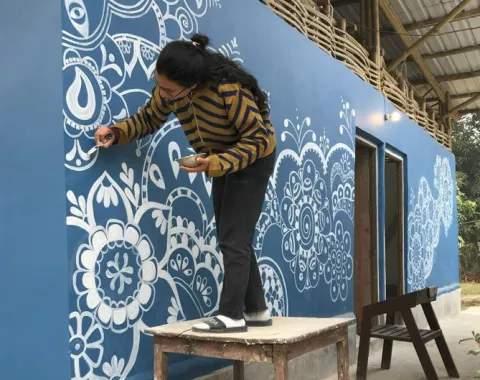 Eine Person bemalt die blaue Außenwand einer Schule in Indien mit weißen, floralen Mustern. Sie steht auf einem Holztisch, um die oberen Bereiche zu erreichen. Das Gebäude hat ein Wellblechdach und ist Teil eines Bildungsprojekts.