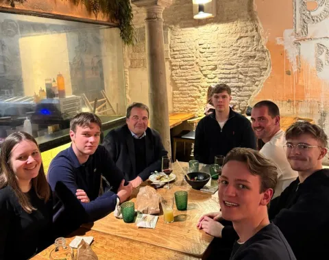 Eine Gruppe von sieben Personen sitzt an einem Tisch in einem Restaurant. Sie lächeln in die Kamera. Im Hintergrund sind eine gemauerte Wand und Pflanzen zu sehen.