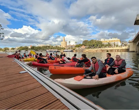 Studierende in Kanus auf einem Fluss in Sevilla, mit Schwimmwesten, vor einer malerischen Stadtlandschaft und bewölktem Himmel.