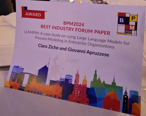 Ein Tischaufsteller mit der Aufschrift "BPM2024 Best Industry Forum Paper" und dem Titel der ausgezeichneten Masterarbeit von Clara Ziche und Giovanni Apruzzese. Im Hintergrund sind stilisierte Skyline-Elemente zu sehen.