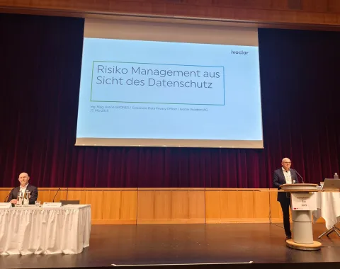 Ein Redner steht an einem Podium auf einer Bühne und hält eine Präsentation mit dem Titel "Risiko Management aus Sicht des Datenschutzes". Ein weiterer Teilnehmer sitzt links am Tisch. Der Hintergrund ist ein roter Vorhang.