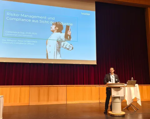 Ein Redner steht auf einer Bühne vor einem großen Bildschirm, der eine Präsentation mit dem Titel „Risiko-Management und Compliance aus Sicht der Industrie“ zeigt. Der Raum hat Holzwände und einen roten Vorhang. Ein Tisch mit Laptop und Mikrofon steht neben dem Redner.