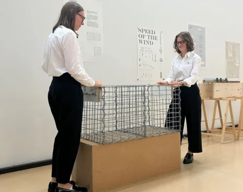Zwei Personen in weißer Kleidung stehen in einem Ausstellungsraum der Universität Liechtenstein. Sie betrachten ein Modell aus Drahtgeflecht auf einem Holzsockel. Im Hintergrund sind Poster mit Architekturzeichnungen zu sehen.