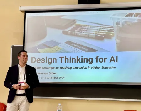 Ein Mann steht vor einer Leinwand, auf der eine Präsentation mit dem Titel "Design Thinking for AI" gezeigt wird. Der Raum wirkt wie ein Seminarraum, und es sind einige Stifte und Notizen auf der Leinwand zu sehen.