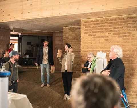 Eine Gruppe von Personen steht in einem Raum mit Wänden aus Stampflehm und Holz. Eine Person hält ein Präsentationsobjekt und spricht zu den anderen. Die Szene zeigt eine Diskussion über nachhaltige Baumaterialien bei einem Symposium der Liechtenstein School of Architecture.