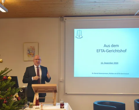 Ein Redner steht neben einem Pult und präsentiert vor einer Leinwand mit der Aufschrift „Aus dem EFTA-Gerichtshof“ bei einer Veranstaltung an der Universität Liechtenstein. Links im Bild ist ein geschmückter Weihnachtsbaum zu sehen.