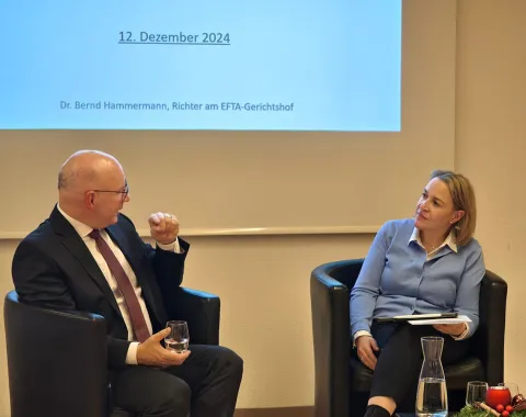 Zwei Personen sitzen in Sesseln vor einer Leinwand, die das Datum "12. Dezember 2024" zeigt. Eine Person hält ein Glas Wasser, die andere hat ein Notizbuch auf dem Schoß. Im Vordergrund steht eine Karaffe auf einem Tisch mit weihnachtlicher Dekoration.