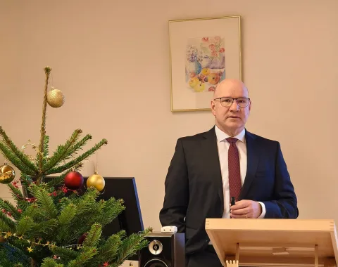 Ein Mann in Anzug und Krawatte steht an einem Rednerpult in einem festlich dekorierten Raum mit einem Weihnachtsbaum. Im Hintergrund hängt ein Bild an der Wand.