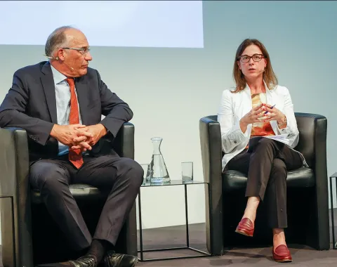 Zwei Personen sitzen auf einer Bühne in schwarzen Sesseln. Die Person links trägt einen dunklen Anzug und eine rote Krawatte, die Person rechts trägt eine weiße Jacke und rote Schuhe. Zwischen ihnen stehen Wasserkaraffen und Gläser.