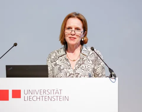 Es ist eine Frau zu sehen, welche vor einem Redepult mit dem Logo der Universität Liechtenstein steht.