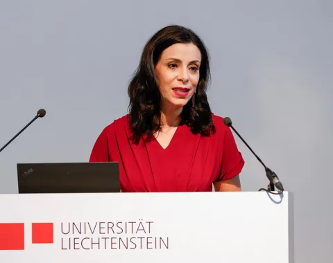 Eine Frau in rotem Kleid spricht an einem Rednerpult mit dem Logo der Universität Liechtenstein. Sie hält eine Rede vor einem Mikrofon.