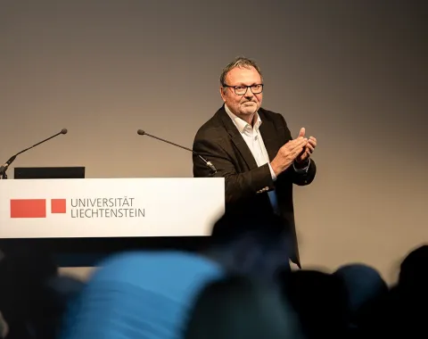 Ein Mann steht an einem Rednerpult der Universität Liechtenstein und applaudiert. Im Hintergrund sind Mikrofone sichtbar.
