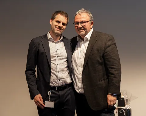 Zwei Männer stehen nebeneinander und lächeln in die Kamera. Sie tragen Anzüge und befinden sich in einem Innenraum mit neutralem Hintergrund.