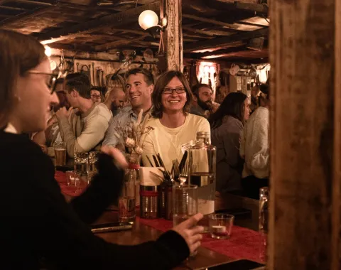Eine Gruppe von Menschen sitzt in einer gemütlichen, rustikalen Bar. Im Vordergrund unterhalten sich Personen an einem Tisch, während im Hintergrund weitere Gäste lächeln und die Atmosphäre genießen. Die Szene wirkt lebhaft und einladend.