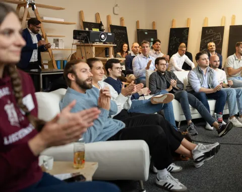 Eine Gruppe von Menschen sitzt in einem Raum und applaudiert. Im Hintergrund sind Tafeln und technische Geräte zu sehen. Die Atmosphäre wirkt konzentriert und feierlich.