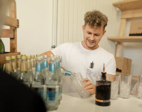 Ein junger Mann schenkt an einem Tisch Getränke ein. Auf dem Tisch stehen mehrere Flaschen und Gläser. Im Hintergrund sind Regale mit weiteren Gegenständen zu sehen.