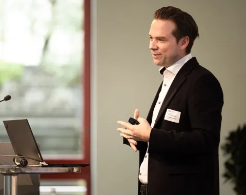 Ein Redner im Anzug hält einen Vortrag auf einer Veranstaltung an der Universität Liechtenstein. Im Hintergrund sind ein Fenster und Pflanzen zu sehen.