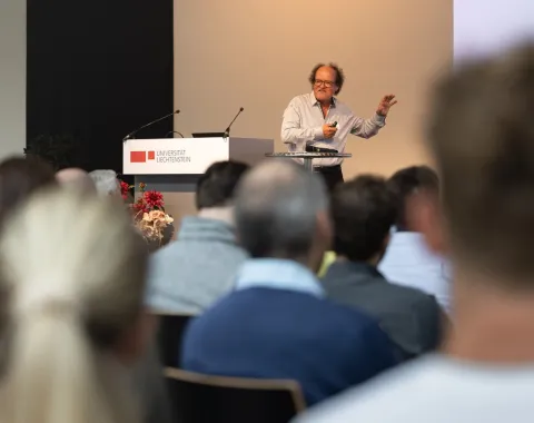 Ein Redner steht an einem Podium und spricht zu einem Publikum bei einer Veranstaltung an der Universität Liechtenstein. Im Hintergrund ist das Logo der Universität sichtbar.