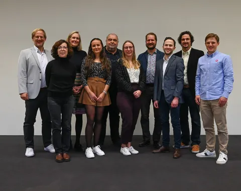 Eine Gruppe von zehn Personen steht lächelnd nebeneinander vor einem neutralen Hintergrund. Sie tragen unterschiedliche, teils formelle, teils legere Kleidung. Das Bild vermittelt eine freundliche und professionelle Atmosphäre.