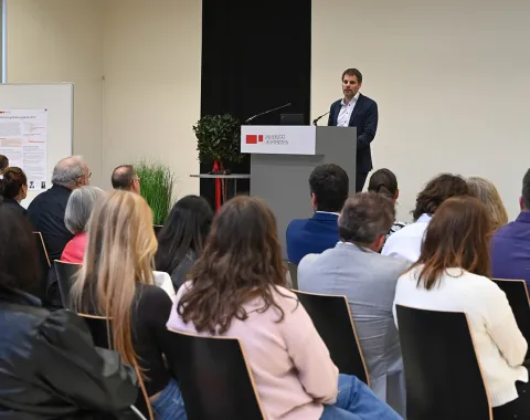 Rektor Christian Frommelt hält eine Rede am Rednerpult vor einem sitzenden Publikum in einem Konferenzraum. Im Hintergrund ist ein Poster sichtbar.