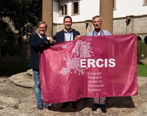 Drei Personen halten ein Banner mit der Aufschrift "ERCIS - European Research Center for Information Systems" in einem sonnigen Innenhof. Im Hintergrund sind historische Gebäude und Säulen zu sehen.