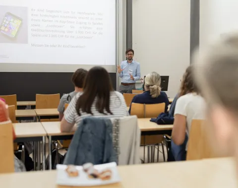 Ein Dozent hält einen Vortrag in einem Seminarraum vor mehreren Erwachsenen. Die Zuhörer sitzen an Tischen und schauen zur Präsentation an der Leinwand.
