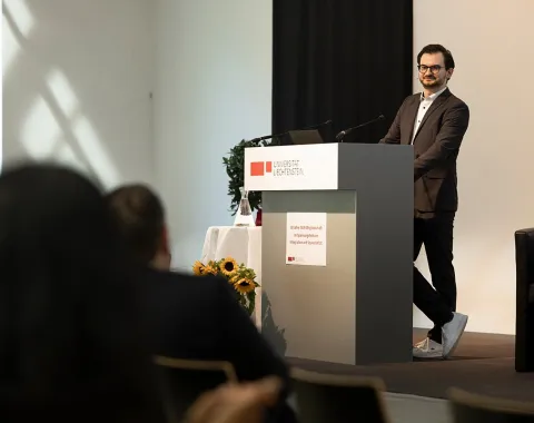 Ein Mann steht an einem Rednerpult in einem Konferenzraum der Universität Liechtenstein. Er trägt einen Anzug und spricht zu einem Publikum. Im Hintergrund sind Sonnenblumen und ein Fenster zu sehen.