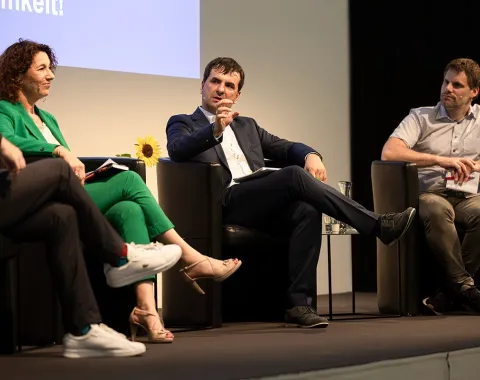 Podiumsdiskussion an der Universität Liechtenstein: Drei Personen sitzen auf einer Bühne in schwarzen Sesseln und diskutieren angeregt. Eine Frau in grünem Anzug und zwei Männer in formeller Kleidung sind zu sehen. Im Hintergrund ist ein Teil einer Leinwand mit Text sichtbar.