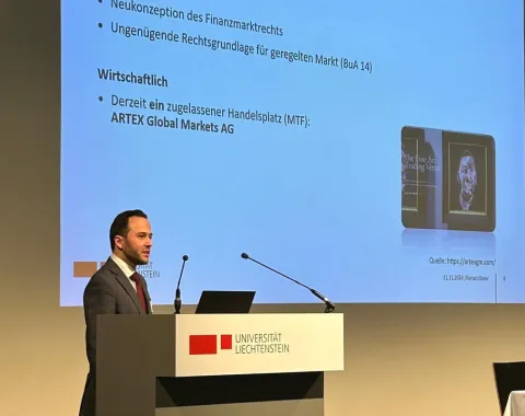 Ein Vortragender steht an einem Rednerpult der Universität Liechtenstein und präsentiert eine Folie mit dem Titel "Hintergrund". Die Folie enthält Informationen zur rechtlichen und wirtschaftlichen Neukonzeption des Finanzmarktrechts.