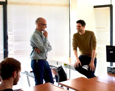 Zwei Männer in einem hellen Raum stehen und sitzen lächelnd an einem Tisch, in einem Gespräch vertieft. Im Hintergrund sind Jalousien zu sehen.