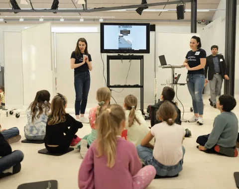 Eine Gruppe von Kindern sitzt auf dem Boden und hört zwei Frauen zu, die vor einem Bildschirm präsentieren. Die Frauen tragen dunkelblaue T-Shirts und Jeans. Im Hintergrund ist ein Mann zu sehen. Der Raum ist hell und modern gestaltet.