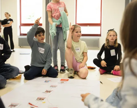 Kinder sitzen in einem Kreis auf dem Boden und nehmen an einem interaktiven Workshop teil. Einige Kinder heben die Hand, um Fragen zu beantworten. Vor ihnen liegt ein großes Papier mit Diagrammen. Die Szene findet in einem hellen Raum mit großen Fenstern statt.