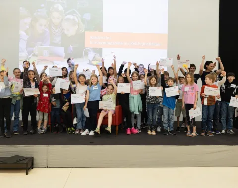 Gruppe von Kindern auf einer Bühne, die stolz Zertifikate hochhalten, im Rahmen der Kinder-Uni an der Universität Liechtenstein. Im Hintergrund ein Bildschirm mit der Aufschrift „Entdeckungsreise ins Reich der Regeln“.